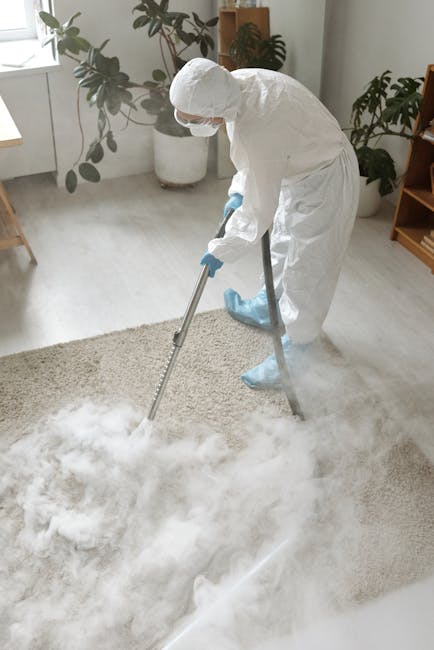 A professional cleaner from Surrey Quays Carpet Cleaners engaged in deep cleaning a beige, textured carpet in a residential living room. The cleaner is dressed in full protective gear, including a whi